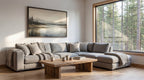 a cozy living room with a large window overlooking a forest, allowing natural light to fill the space. The room features a gray sectional sofa, a wooden coffee table, and a framed landscape painting on the wall.