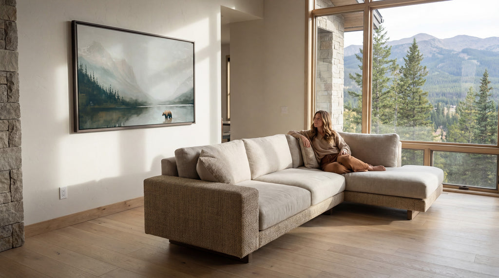 A woman sits on a beige sectional sofa in a spacious living room, gazing out a large window overlooking a scenic mountain landscape.