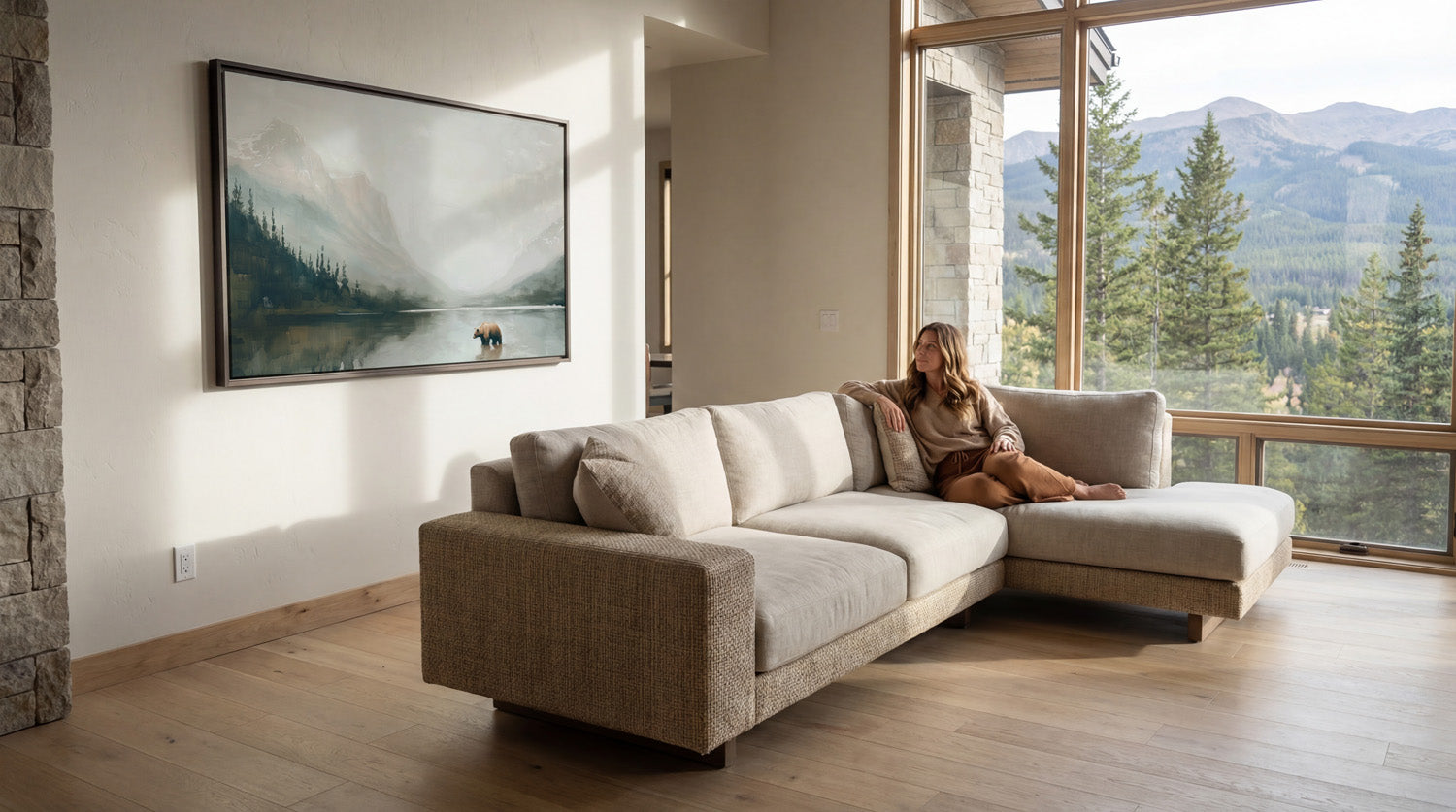 A woman sits on a beige sectional sofa in a spacious living room, gazing out a large window overlooking a scenic mountain landscape.