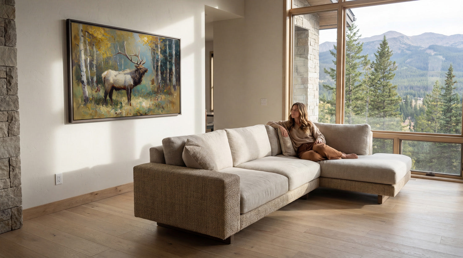 A woman sits on a beige sectional sofa in a spacious living room, with a large window offering a scenic view of a forest and mountains. A framed painting of a deer hangs on the wall above the sofa.