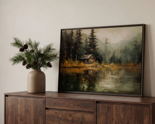 a wooden cabinet with a framed painting of a cabin in a forest, placed on top of it. Next to the painting, there is a vase with pine branches and pine cones.