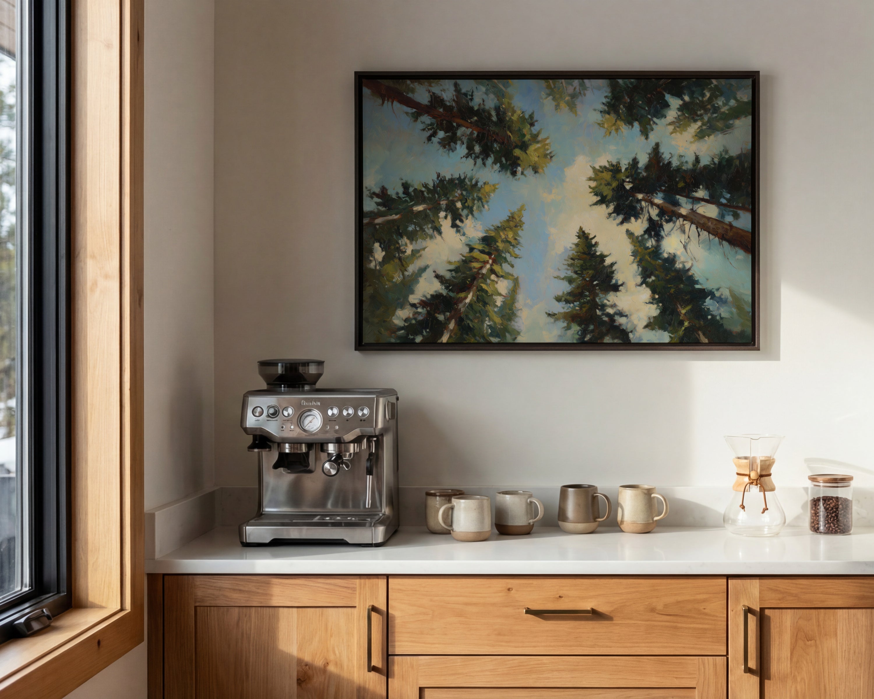 a kitchen counter with a coffee machine, coffee cups, and a framed painting of a forest scene above it.