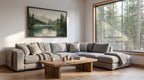 a cozy living room with a large window overlooking a forest, allowing natural light to fill the space. The room features a gray sectional sofa, a wooden coffee table, and a framed landscape painting on the wall.