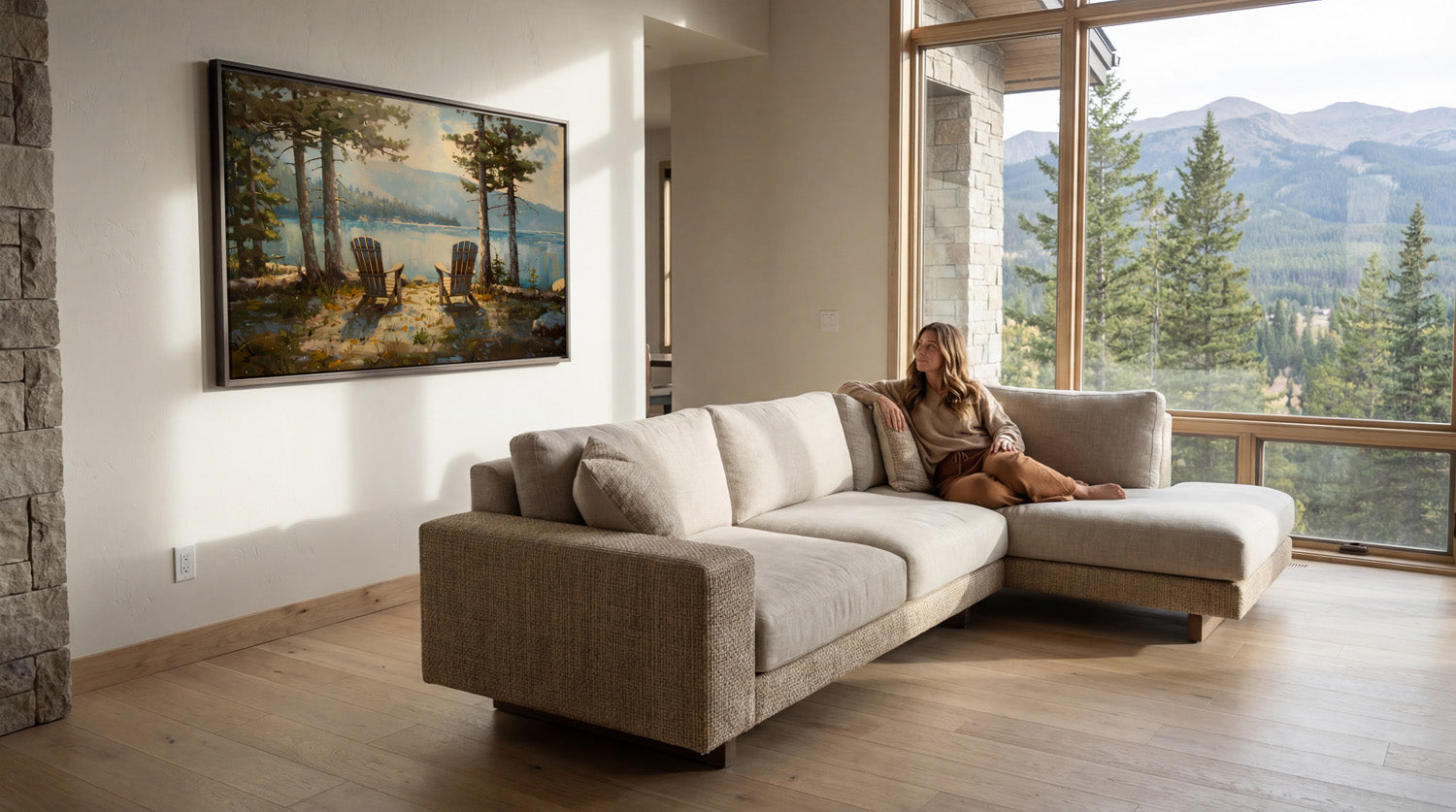 A woman sits on a beige sectional sofa in a spacious living room, with a large window offering a scenic view of a forested mountain range.