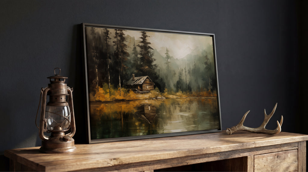 a painting of a cabin surrounded by trees, with a lake in the foreground. The painting is displayed on a wooden table, and there is a lantern and antlers next to it.