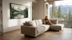 A woman sits on a beige sectional sofa in a spacious living room, gazing out a large window overlooking a scenic mountain landscape.