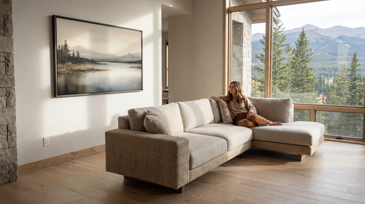 A woman sits on a beige sectional sofa in a spacious living room, gazing out a large window overlooking a scenic mountain landscape.