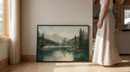 A woman in a long white skirt stands next to a large framed painting depicting a serene landscape with a lake and mountains.