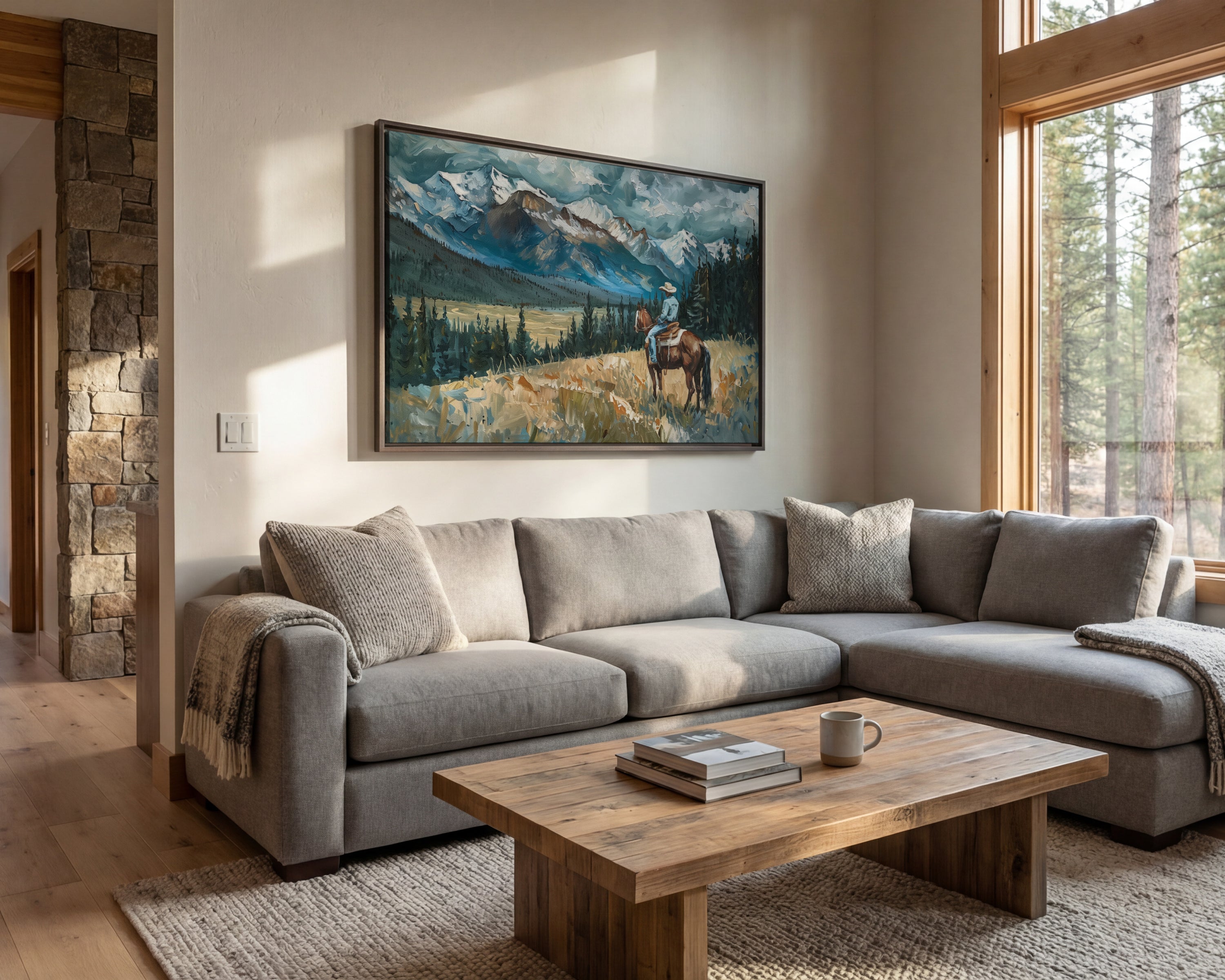 a cozy living room with a large painting of a mountain landscape hanging on the wall above a gray sectional sofa. The sofa is positioned in front of a wooden coffee table, which has a stack of books and a mug on it. The room is illuminated by natural light coming through a large window, and the overall atmosphere is warm and inviting.
