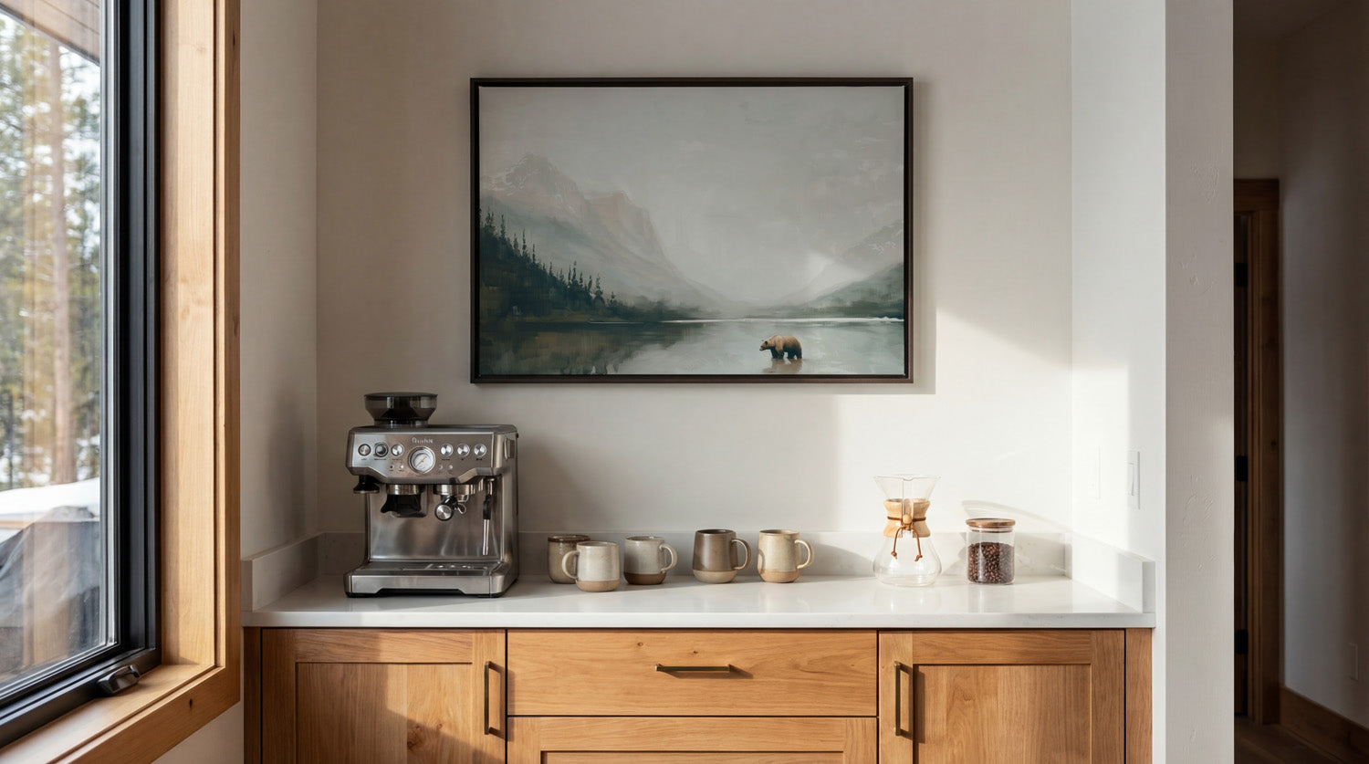 a cozy kitchen scene with a framed landscape painting hanging on the wall above a white countertop. On the countertop, there is a coffee maker and two mugs, suggesting a space for enjoying a warm beverage.