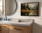 a bathroom with a sink, a mirror, and a framed painting of a cabin in a forest. The painting is hanging on the wall above the sink, which has a wooden cabinet below it. The sink has a black faucet, and there are some towels on the counter.