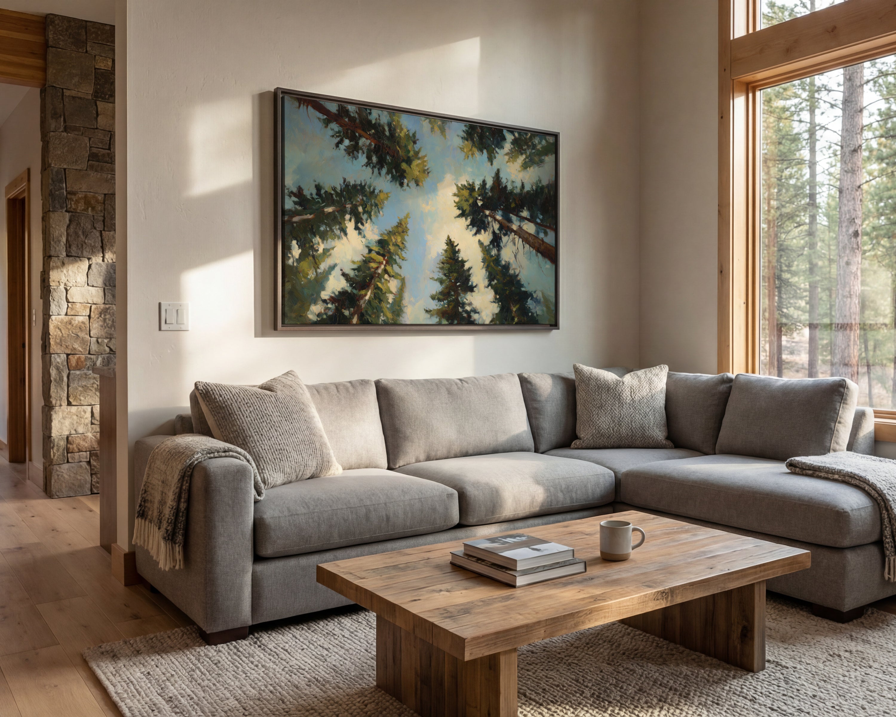a cozy living room with a large window overlooking a wooded area, allowing natural light to fill the space. The room features a gray sectional sofa, a wooden coffee table, and a framed landscape painting on the wall.