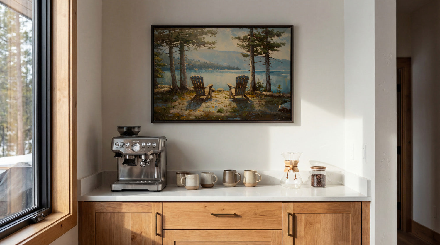 a cozy kitchen scene with a framed painting of a serene lake and two chairs, a coffee machine, and various kitchen accessories on a white countertop.