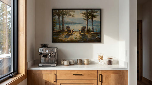 a cozy kitchen scene with a framed painting of a serene lake and two chairs, a coffee machine, and various kitchen accessories on a white countertop.