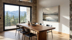 a modern dining room with a large wooden table and chairs, a framed landscape painting on the wall, and a view of mountains through a large window.