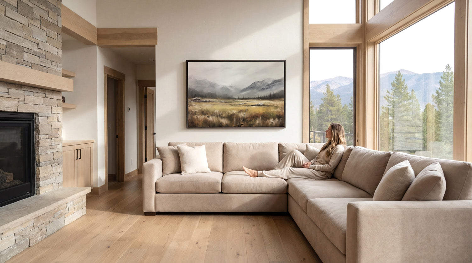 A woman sits on a large beige sectional sofa in a spacious living room, with a fireplace and large windows offering a scenic view of mountains.