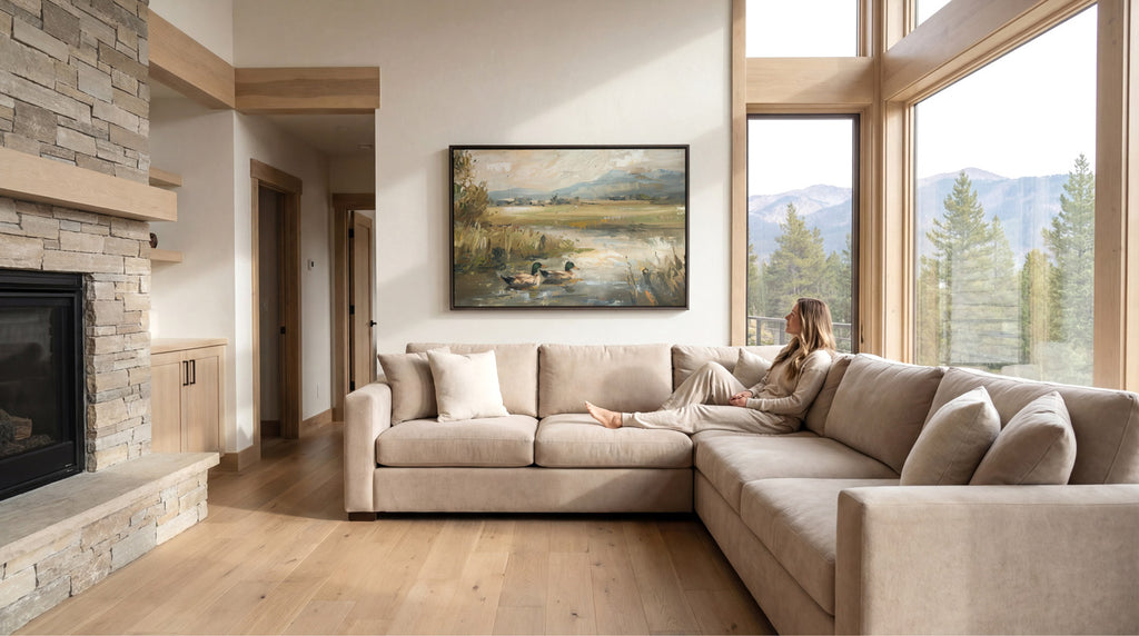 A woman sits on a beige sectional sofa in a spacious living room, with a large painting of a landscape hanging on the wall above her. The room has a rustic, cabin-like aesthetic with a stone fireplace and large windows offering a scenic view of the mountains outside.