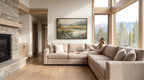 A woman sits on a beige sectional sofa in a spacious living room, with a large painting of a landscape hanging on the wall above her. The room has a rustic, cabin-like aesthetic with a stone fireplace and large windows offering a scenic view of the mountains outside.