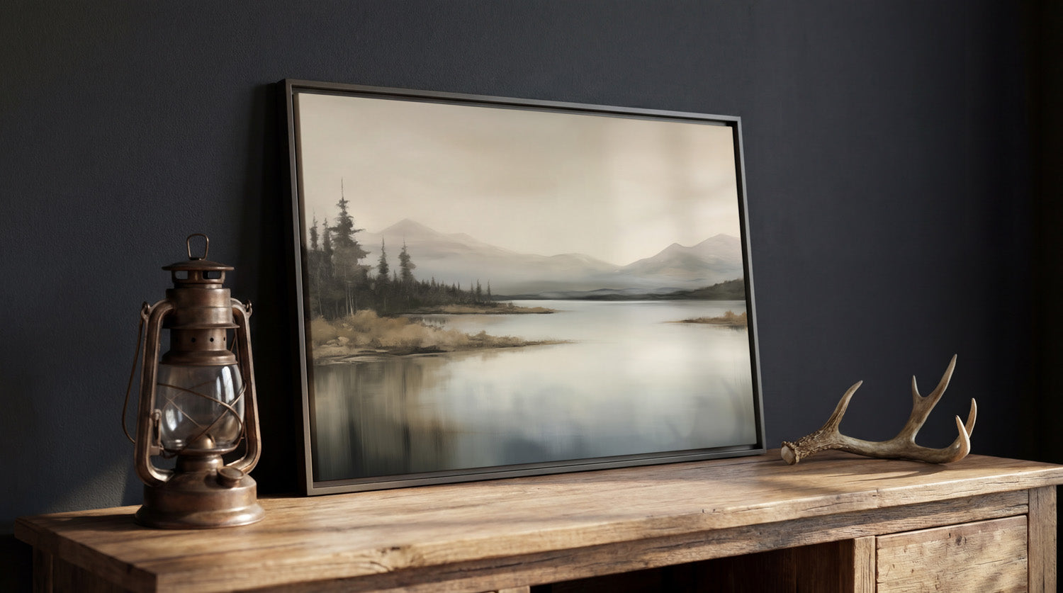 a framed landscape painting depicting a serene mountain lake scene, with a lantern and antler decoration placed on a wooden table.