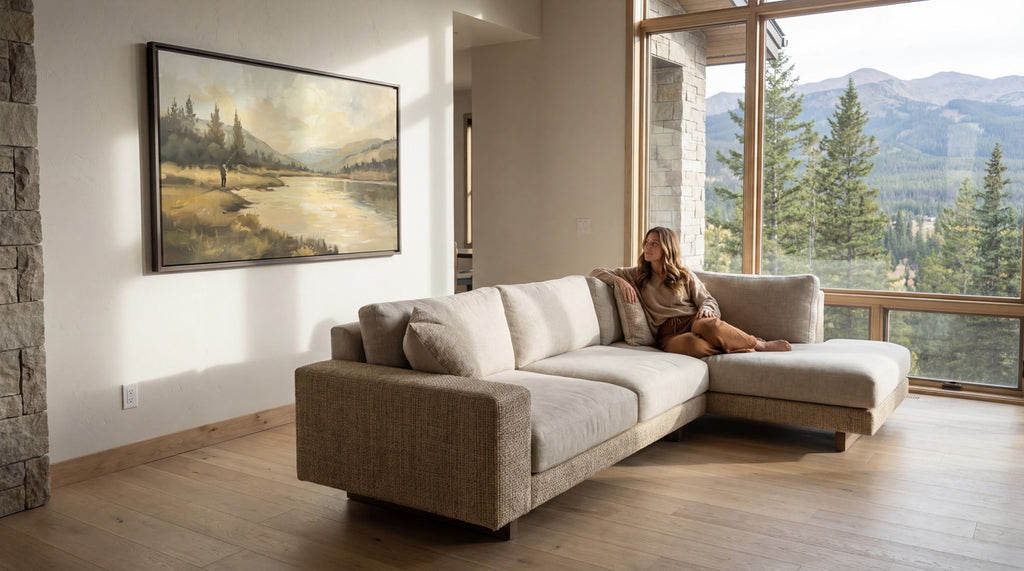 A woman sits on a beige sectional sofa in a spacious living room, with a large window offering a scenic view of a mountain landscape.