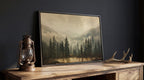 a framed landscape painting depicting a serene forest scene with pine trees and a lake, accompanied by a lantern and antlers on a wooden table.