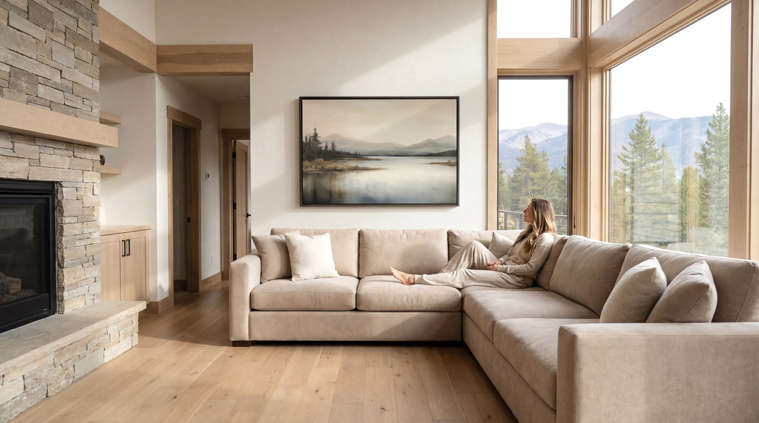 A woman sits on a large beige sectional sofa in a spacious living room, with a fireplace and large windows offering a scenic view of mountains.