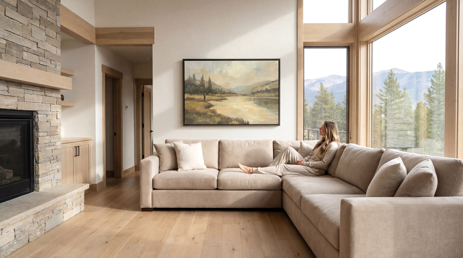 A woman sits on a large beige sectional sofa in a spacious living room, with a fireplace and large windows offering a scenic view of the mountains outside.