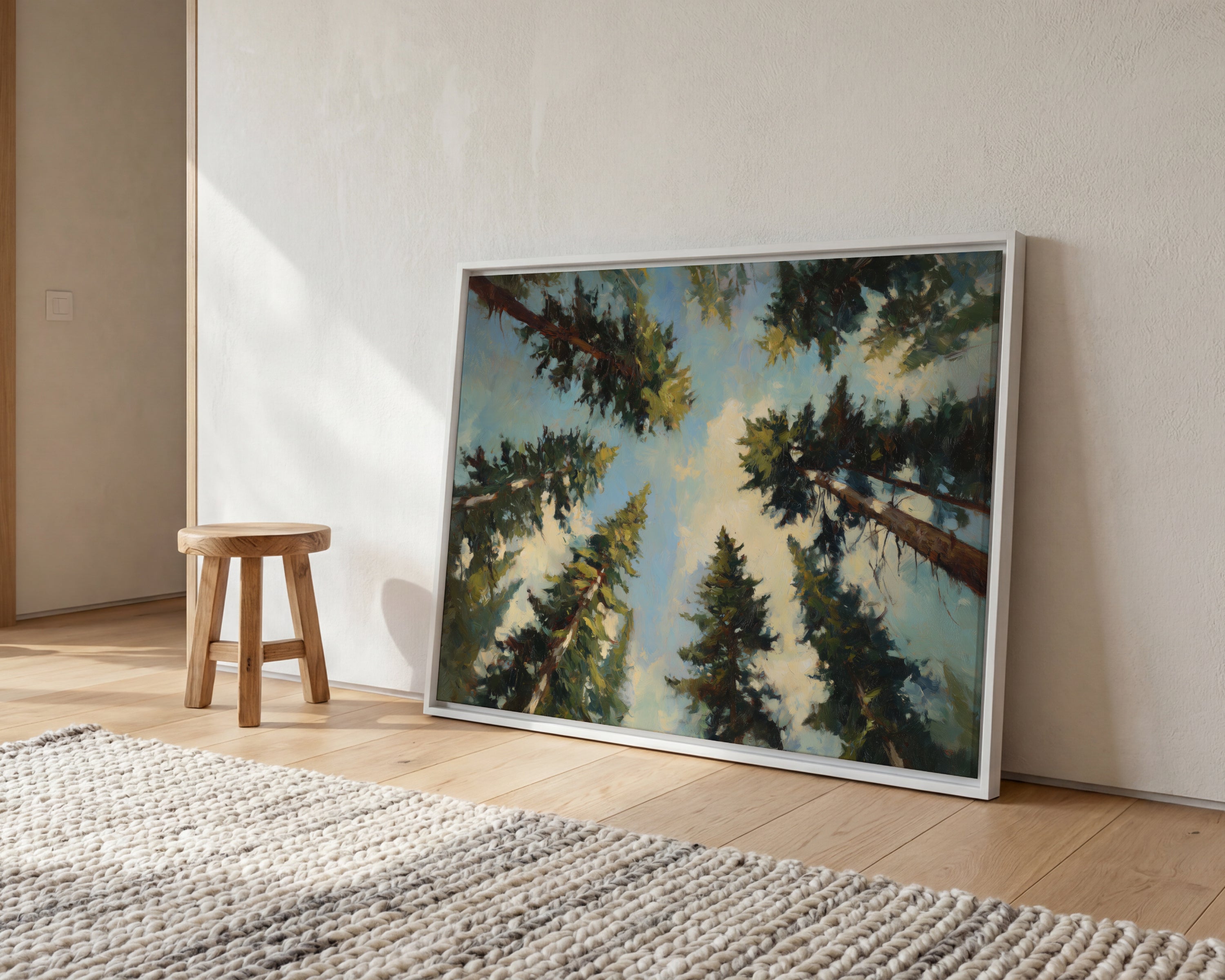 a large painting of a forest scene with tall trees reaching towards a bright blue sky, displayed on a white wall in a room with a wooden stool and a woven rug.