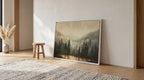 a large framed landscape painting depicting a forest scene with a lake in the background, placed on a wooden floor in a room with a rug and a small stool.