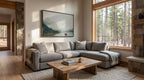 a cozy living room with a large window overlooking a forest, a gray sectional sofa, a wooden coffee table, and a framed landscape painting on the wall.